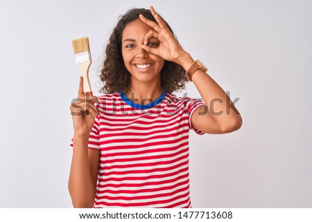 Young brazilian painter woman holding brush standing over isolated white background with happy face smiling doing ok sign with hand on eye looking through fingers