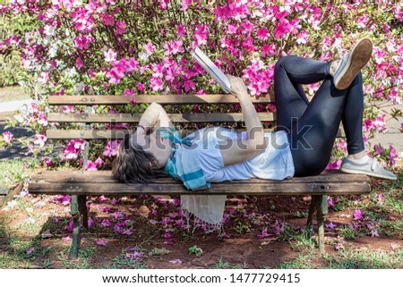 40 years old woman model lying on wooden bench reading a book in the park in front of an azaleas bush - 40-year-old or middle-aged woman reading a book in the park