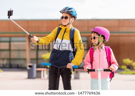 education, childhood and people concept - happy school children in helmets with backpacks riding scooters and taking picture by smartphone on selfie stick outdoors
