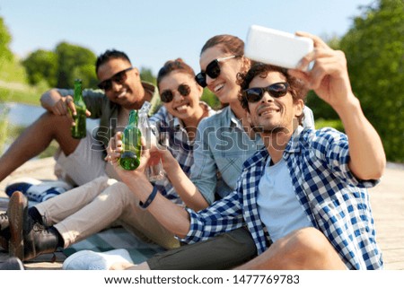 leisure, picnic and technology concept - friends with drinks taking selfie by smartphone on lake pier in summer park