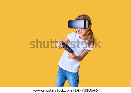 Girl experiencing VR headset vs joystick game on yellow background. Surprised emotions on her face. Child using a gaming gadget for virtual reality. Futuristic goggles at young age. Virtual technology