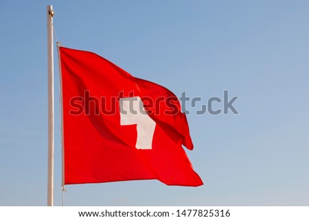 The Flag of Switzerland flutters in the wind against a blue sky during a sunny day.