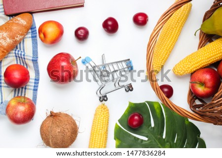 Buying food for a picnic. Mini shopping trolley with fruits and basket on a white background. Monstera leaf.