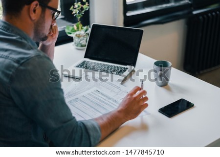 Serious pensive thoughtful focused young casual businessman or entrepreneur in office filling income tax return papers documents for IRS