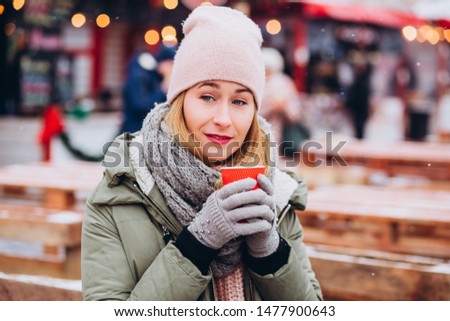 Girl in a pink hat and a knitted woolen scarf drinks cocoa at the Christmas market. woman with a red glass on a city holiday. girl basks in hot tea on the background of the Christmas market.