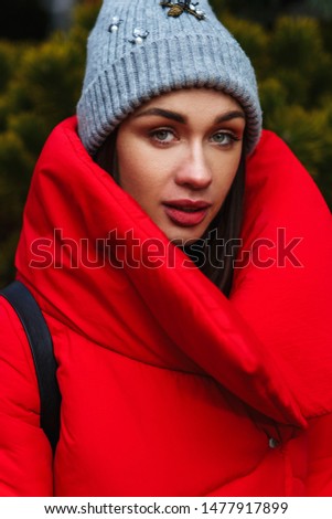 Portrait of young attractive woman in a red down jacket standing on the winter street and looking at camera. Outdoor photo of dreamy woman in warm clothes. City lifestyle