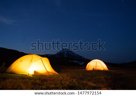 Night photo of the camping site in Kamchatka, Russia. Yellow tents with light inside in the darkness. Stars in the sky. Silhouette of symmetrical cone of Vilyuchik volcano in dark blue  background.