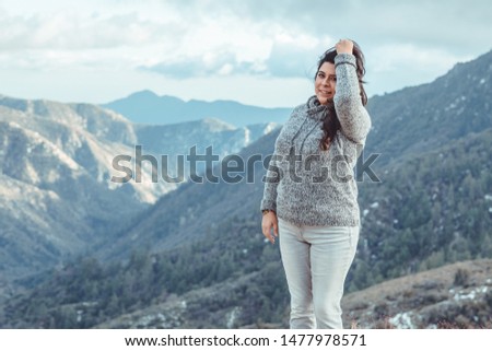 A young woman with a happy smile and facial expression while posing for a picture. playing with her hair also. background is a mountain landscape during the winter time with cool blue tone colors. 