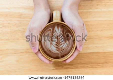 Selective focus.Woman 's hand holding a cup of hot latte art coffee (leaf pattern) on wooden table background at cafe shop.Concept : Holiday ,Day off work ,Weekend and Sharing.