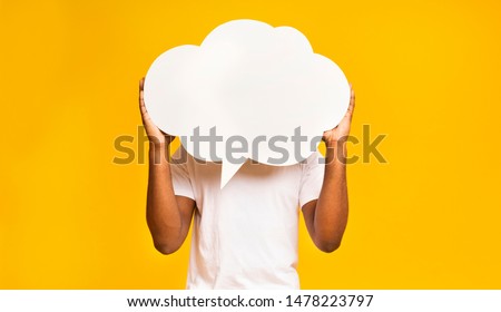 Unrecognizable black man holding an empty speech bubble in front of his head, yellow studio background, empty space Royalty-Free Stock Photo #1478223797