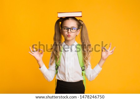 Back To School Concept. Cute Nerdy Schoolgirl Meditating With Book On Head On Yellow Studio Background. Free Space