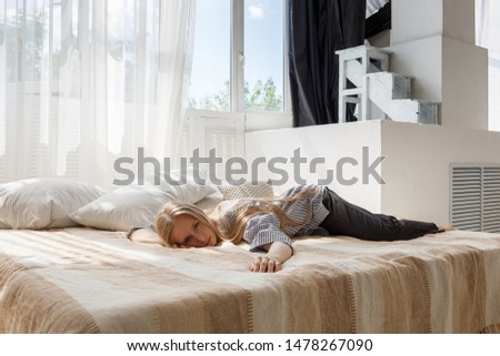cute blonde girl with long hair in a striped shirt and black pants lies in bed at bright sunny studio face down and looks at camera. behind her there are pillows and window with transparent curtain