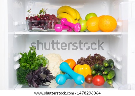 on the shelves of a white refrigerator, a stock of fresh vegetables, fruits, berries, as well as pink and blue dumbbells for sports