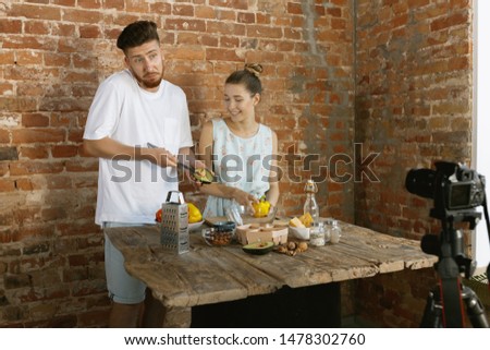 Vlogger and blogger freelance job, food concept. Young caucasian couple cooking together and recording live video for vlog and social media with professional camera against brick wall in their kitchen