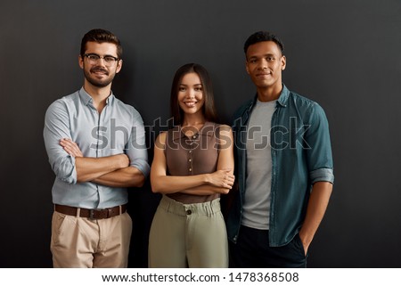 We are great team. Group of three multicultural cheerful young people in casual wear looking at camera with smile while standing against dark background