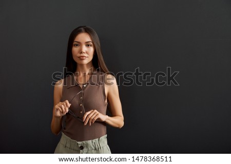 Successful business woman holding eyeglasses and looking at camera while standing against dark background