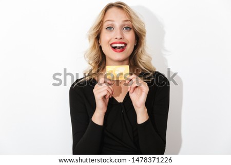 Portrait of an attractive excited blonde woman dressed in black dress standing isolated over white background, holding plastic credit card
