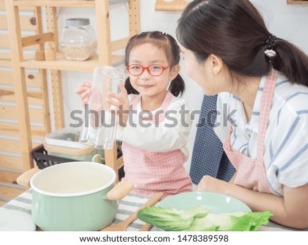 Asian woman wearing pink apron teaching smiley little girl wearing red grasses cooking spaghetti food with vegetable and pot on wooden table in kitchen. soft tone picture