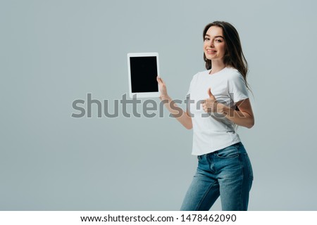 smiling beautiful girl in white t-shirt showing digital tablet with blank screen and thumb up isolated on grey