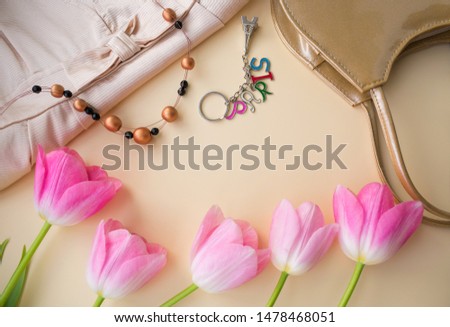A composition of five buds of pink flowers, fragments of a handbag, dress, necklace and keychain similar to the Eiffel Tower with the inscription Paris. Top view on a beige background