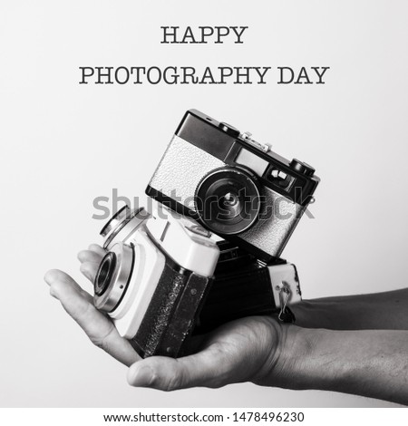 closeup of the hand of a caucasian man holding some retro film cameras and the text happy photography day, in black and white