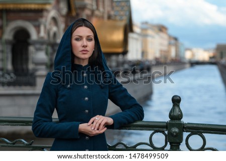 Poetic brown hair brunette sister beautiful woman girl vintage classic coat blue dress look camera green eyes hands together street old town outdoor blue sky river cathedral channel scenic city view Royalty-Free Stock Photo #1478499509