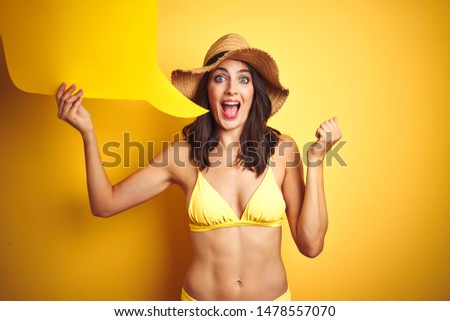 Beautiful woman wearing yellow bikini and holding talking balloon over isolated yellow background screaming proud and celebrating victory and success very excited, cheering emotion