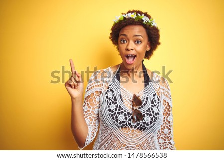 Young african american woman with afro hair wearing flowers crown over yellow isolated background pointing finger up with successful idea. Exited and happy. Number one.