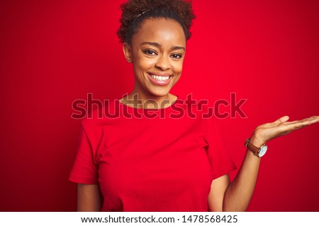 Young beautiful african american woman with afro hair over isolated red background smiling cheerful presenting and pointing with palm of hand looking at the camera.