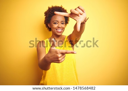 Beauitul african american woman wearing summer t-shirt over isolated yellow background smiling making frame with hands and fingers with happy face. Creativity and photography concept.