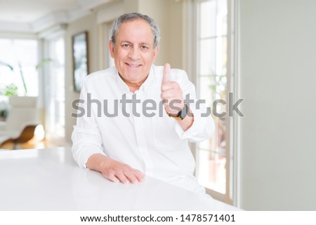 Handsome senior man at home doing happy thumbs up gesture with hand. Approving expression looking at the camera with showing success.