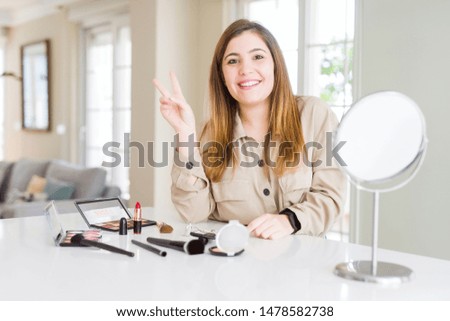 Beautiful young woman using make up cosmetics smiling with happy face winking at the camera doing victory sign. Number two.