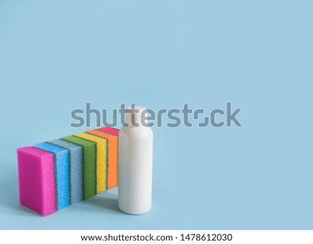 on a blue background are seven rectangular washcloths in a row of sponges for dishes for cleaning the purity of all the colors of the rainbow and next to it is a white plastic bottle with a detergent 