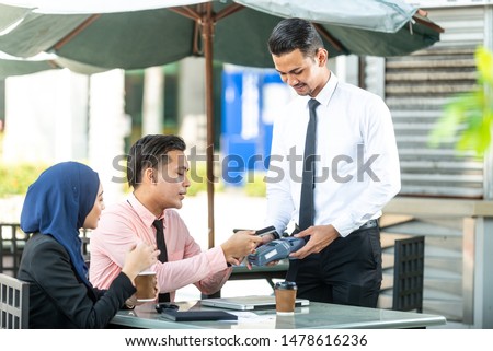 Waiter interacting with Muslim customers using bank terminal to process and acquire mobile payment at a coffee shop on a sunny day. Modern cafe start up small business micropayment concept.