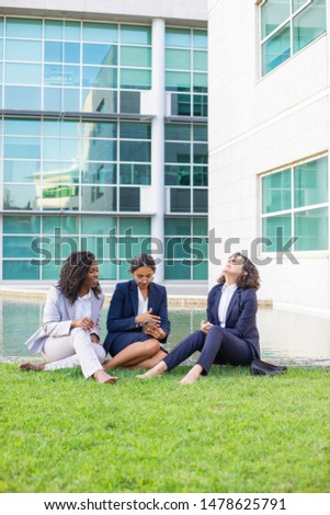 Barefoot smiling businesswomen with smartphone outdoor. Young multiethnic female colleagues sitting on green lawn and using mobile phone. Technology concept