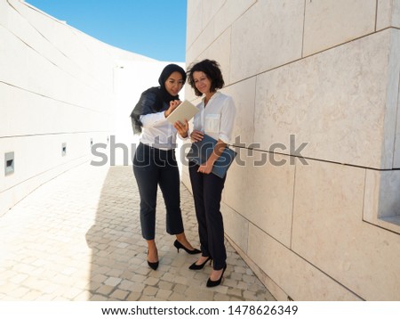 Multicultural managers checking report on tablet. Business women in office suits and hijab standing outside, using tablet and looking at screen together. Digital technology in global business concept