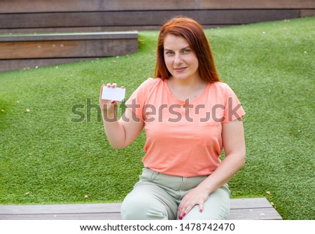 Happy beautiful ginger woman holding white card isolated on green background. Happiness emotion. girl sitting on a bench in the park.