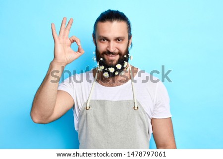 A handsome man with flowers in beard in gray apron showing ok gesture isolated on blue background. isolated blue background. studio shot . body language
