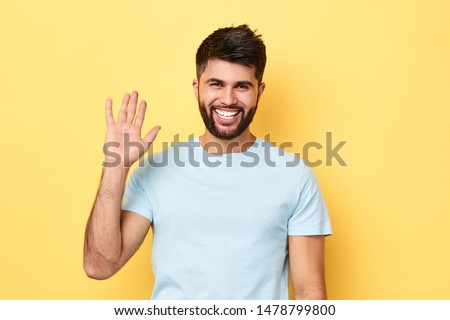 Hello Hi Young cheerful handsome bearded man greeting with open hand, enjoying communication, isolated on yellow background. close up portrait, body language.