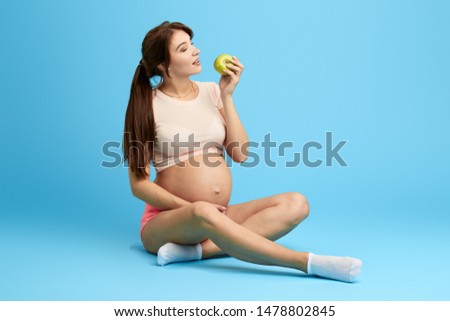 woman with big belly eating green apple, vitamin for baby. full length photo. isolated blue background. studio shot. apple a day keeps the doctor away