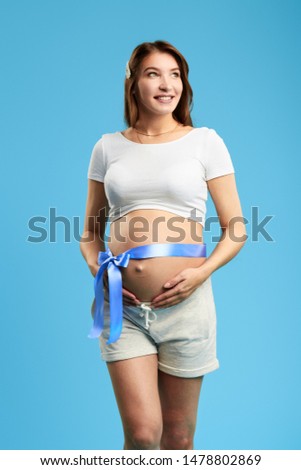 happy beautiful pensive young pregnant woman with blue ribbon around her belly looking away standing over blue background. close up photo. isolated blue background. studio shot.lifestyle