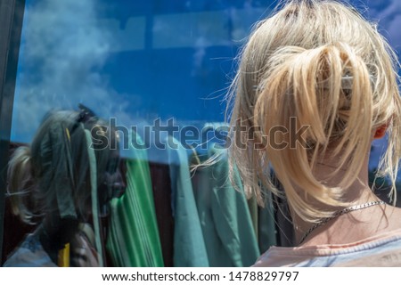 A young blonde girl with a hairpin on her head in a striped T-shirt is looking at shop windows with clothes on a sunny day, a photo from the back