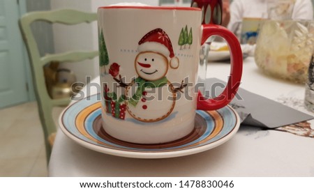 Handcrafted Christmas mug for hot winter drinks. White ceramic cup with red handle and picture of cute snowman in red Santa's hat. Colorful crockery closeup in green kitchen interior. 