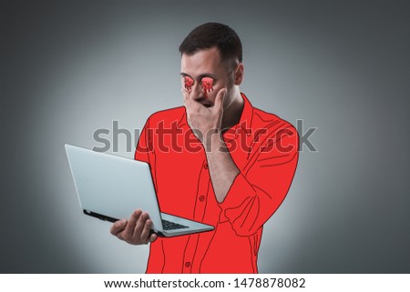 Handsome young man with a cartoon eyes and shirt is holding a laptop, looking at it and acting like he is shocked.