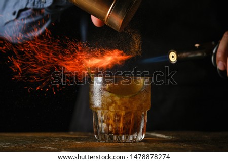 Barman prepares cocktail with
lime  and herbs in transparent glass on bar with alcohol. Uses burner with sparks. Dark background.