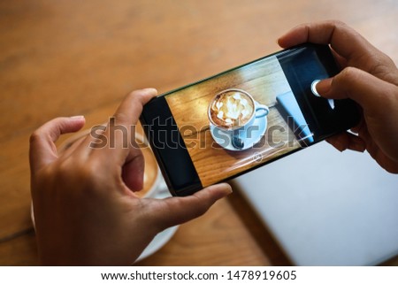 Hand taking a picture of hot latte art coffee cup with art foam on wooden table  at a coffee shop snap and share on social media by smartphone. Web Blogger photo shoot.