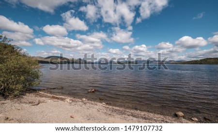 A canoeist paddles across a calm Loch Lomond in Scotland with blue skies and fluffy clouds overhead and hills in the distance