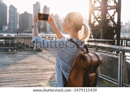 Back view of millennial woman shooting video of New York landscape using smartphone for clicking pictures, positive female tourist testing cellphone camera for taking photos of city in sunrise