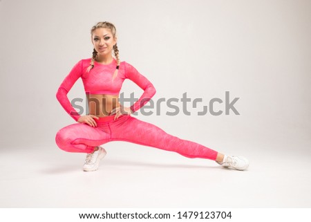 Sport exercises on a grey background, fitness concept. A young woman in comfortable sportswear smiling charmingly and doing wide lunges to the sides with her legs on an isolated grey background.