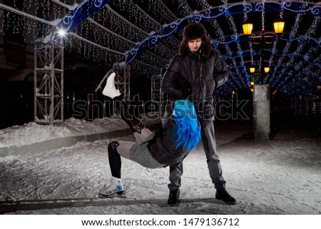 Couple on the city rink in a winter evening. Guy helping a fallen girl with blue hair on the ice in the dark night and twinkles lighting above them